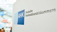 AHK-Logo auf einer Stellwand, im Hintergrund Messegeschehen