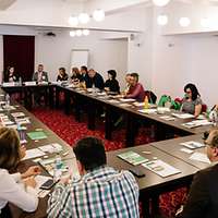 Zahlreiche Männer und Frauen sitzen an einem großen, über Eck gebauten Tisch voller Unterlagen 